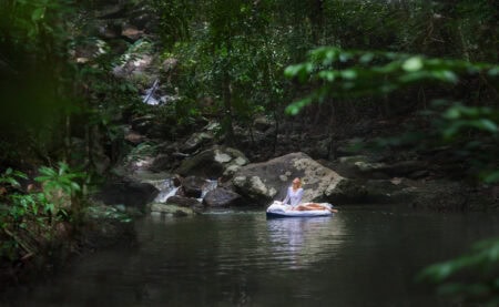 Flowing stream in lush green rainforest with woman meditating on float mattress, peaceful nature scene, outdoor relaxation, natural wellness, tranquil water body, eco-friendly lifestyle, mind-body harmony.